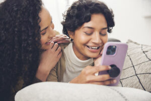 Two women looking at a mobile phone and laughing together on a couch.