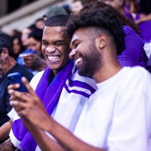Two people at a sports game, looking at a phone and smiling.